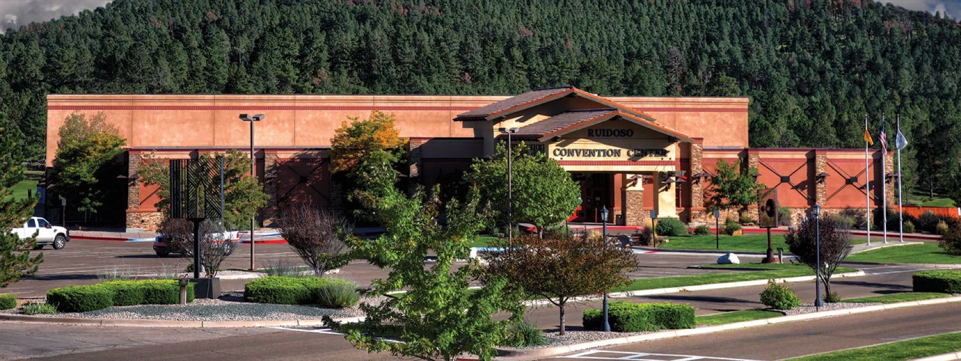 A wide autumn view of the building with flags flying out front and forested mountains rising directly behind it.