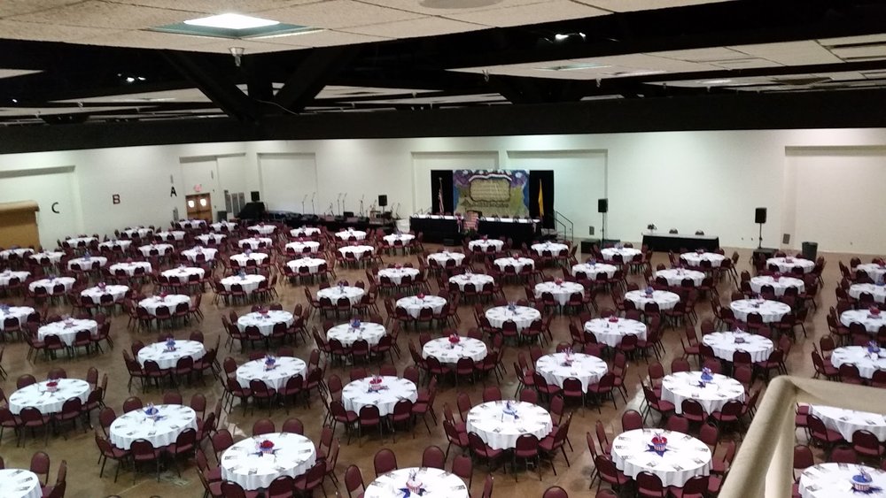 An elevated view from a balcony overlooking the full Exhibit Hall set for a banquet, rows of round tables filling the floor with lettered seating sections marked on the walls.