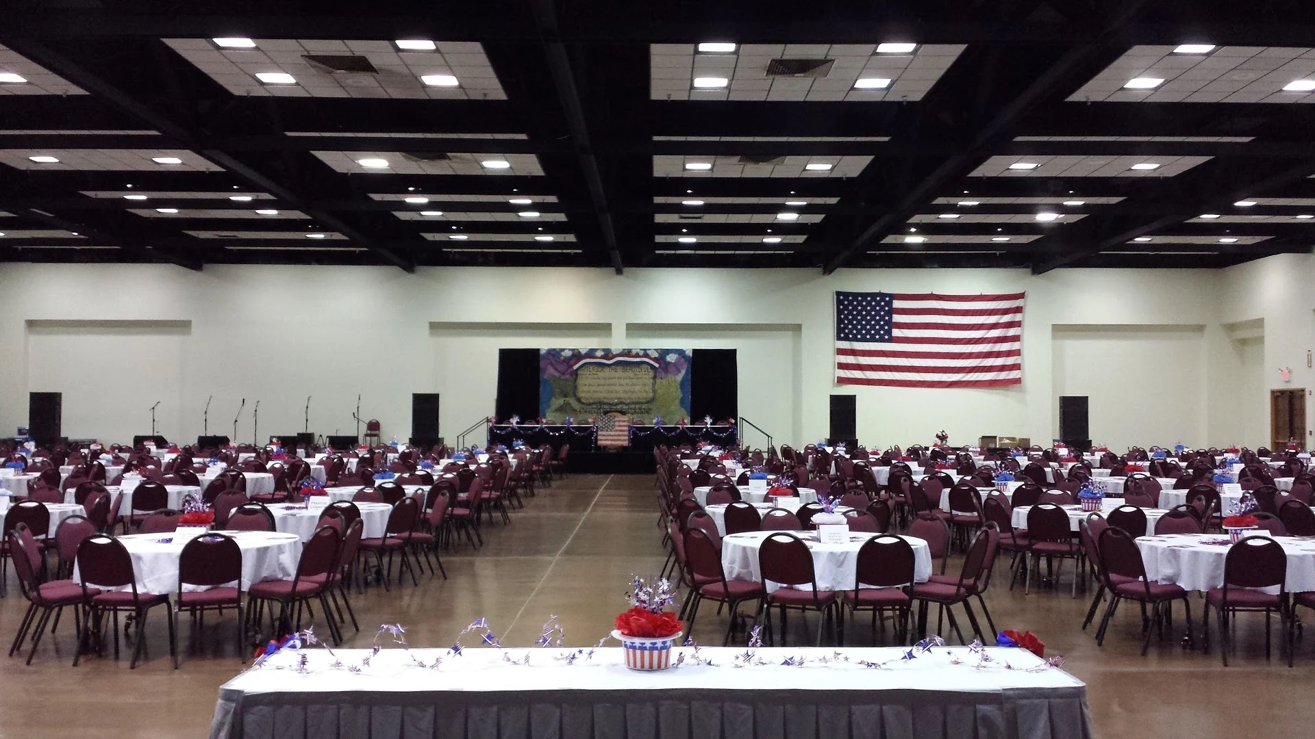 A large hall set for a banquet with rows of round tables in red-and-white linens, a low stage with a painted backdrop, and a large American flag on the side wall.