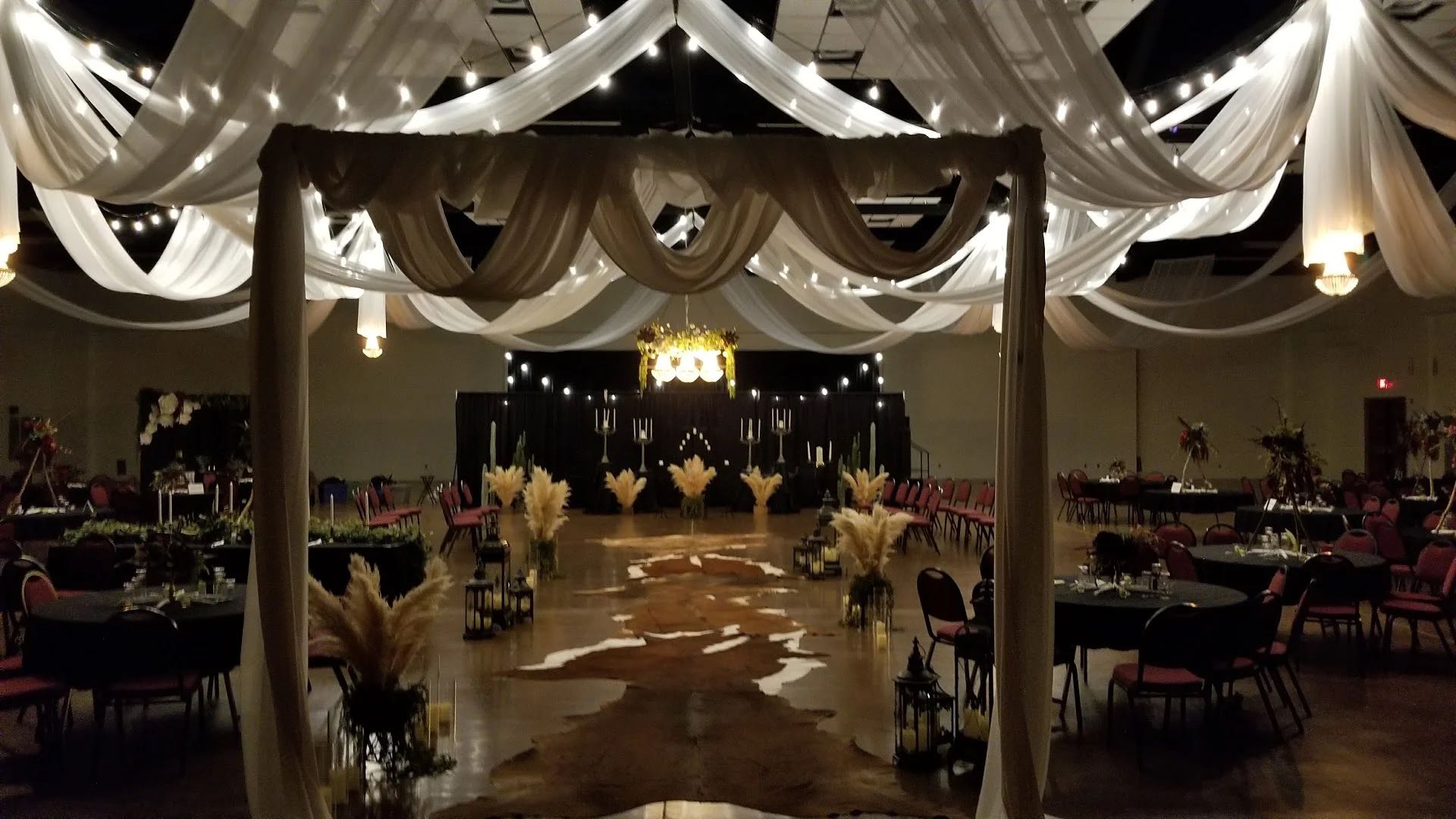 An evening reception in the Exhibit Hall: string lights and draping over a rustic hide-covered aisle, pampas grass and candelabra flanking the walk.