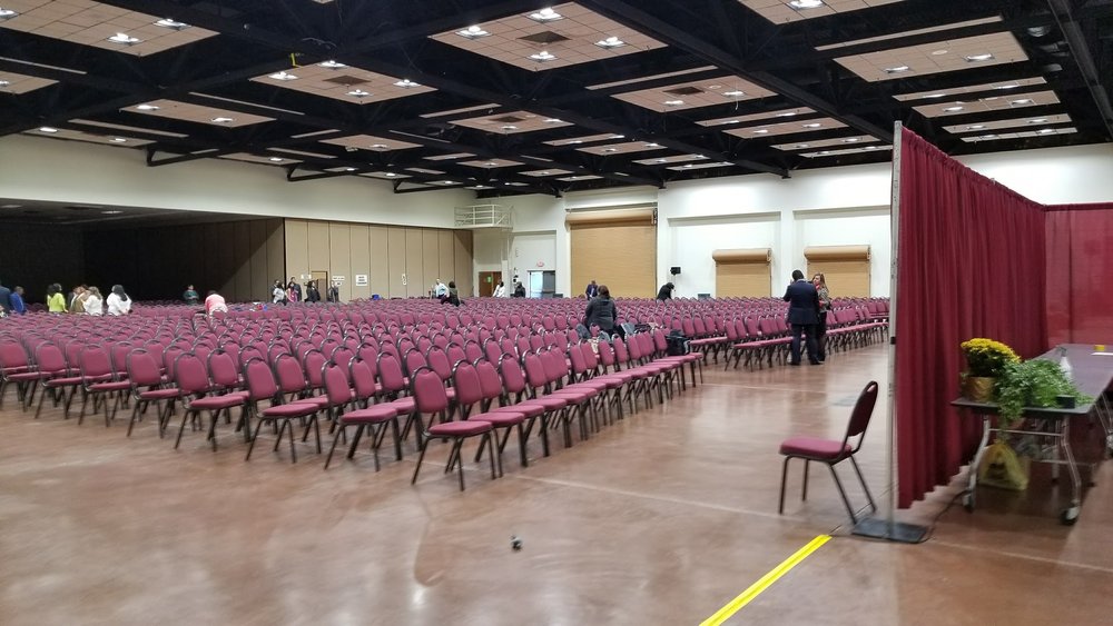 Hundreds of chairs in theater-style rows filling the Exhibit Hall for a ceremony, with a red pipe-and-drape divider being set up to one side.