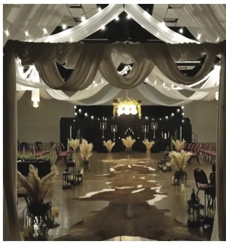 A wedding ceremony aisle inside the Convention Center, framed by a draped fabric arch with string lights, leading to a floral altar.