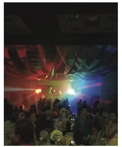 A wedding reception in full swing under colored uplighting and draped fabric ceiling treatment, guests dancing.